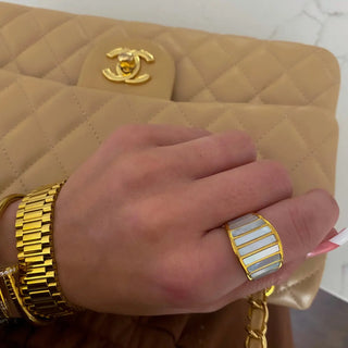 Hand wearing a gold ring and bracelet with a beige quilted handbag featuring a gold logo.