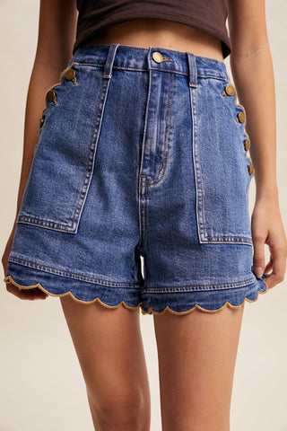 Blue denim shorts with scalloped hem and gold buttons on a beige background