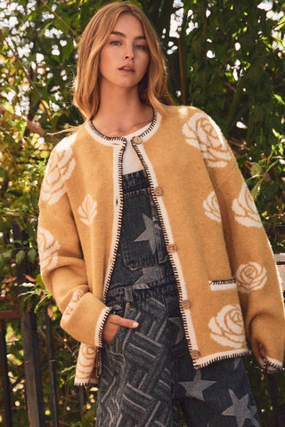 A woman wearing a mustard yellow fuzzy cardigan with cream rose and leaf patterns, styled over denim overalls in an outdoor garden setting.