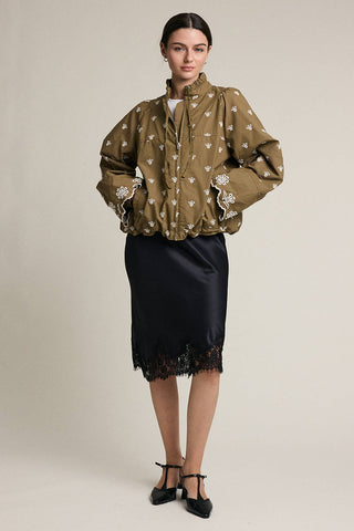 A model stands against a plain beige studio background wearing a cropped olive-brown jacket with white floral embroidery and a black satin midi skirt with a black lace hem, paired with black Mary Jane heels.