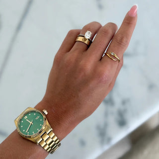 Hand wearing a gold watch with a green face and multiple rings on a blurred background