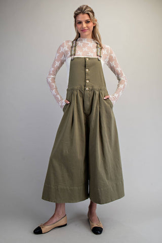 A woman modeling a sage green wide-leg bib jumpsuit with front buttons, worn over a sheer white floral long-sleeve top and paired with beige-and-black flats, against a neutral gray studio background.