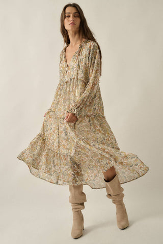 A woman modeling a long-sleeved, tiered midi dress with a beige and yellow floral print, paired with beige suede boots, against a plain beige studio background.