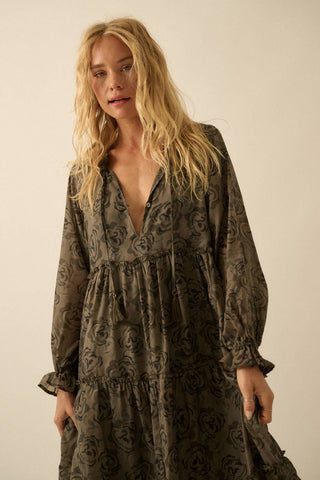 A woman wearing a long-sleeve, tiered midi dress with a dark floral pattern on a muted olive-brown base, posed against a plain beige studio background.
