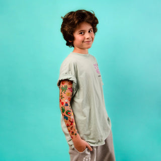 A child wearing a light sage green t-shirt with a colorful cartoon character printed on the sleeve, standing against a solid turquoise background.