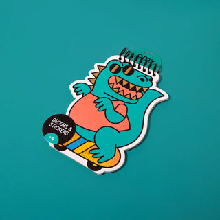 A die-cut sticker featuring a cartoon teal dinosaur wearing sunglasses and a pink shirt while riding a yellow skateboard, shown on a solid teal background. The sticker has a black spiral binding at the top and a black circular label that reads 'DECORS & STICKERS'.