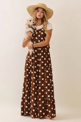 Woman in a brown polka dot dress with a white top, straw hat, and small dog on a plain background