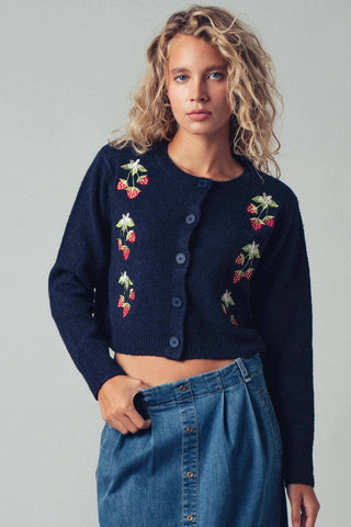 Woman wearing a navy blue cardigan with floral embroidery and blue jeans on a gray background