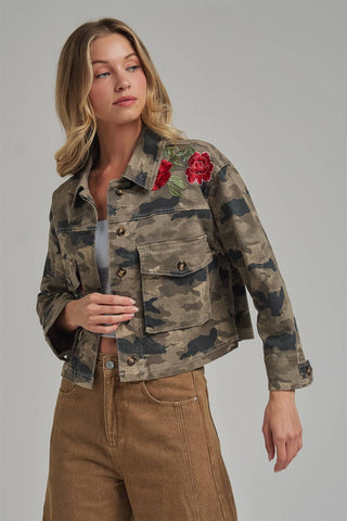 A woman models a cropped camouflage denim jacket featuring red rose embroidery on the chest, paired with brown pants, against a solid gray studio background.