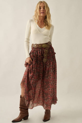 A woman modeling a long, flowy bohemian-style skirt with a red and green floral pattern, a wide brown belt with large decorative medallions, paired with a cream long-sleeve top and brown leather boots, against a plain light beige studio background.