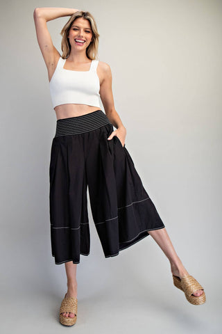 A woman modeling black wide-leg cropped pants with a black and white striped waistband, paired with a white crop top and woven sandals, against a neutral gray studio background.