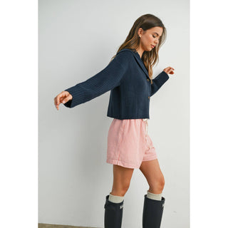 Woman wearing a navy cardigan, pink shorts, and black boots against a white background