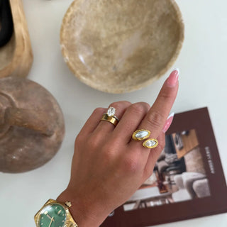 Hand wearing gold rings with a watch, set against a neutral background with decorative items.