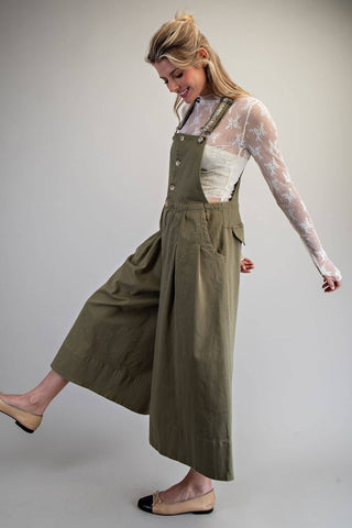 A woman modeling olive green linen wide-leg overall jumpsuit over a white lace long-sleeve top, posing against a neutral light gray studio background.