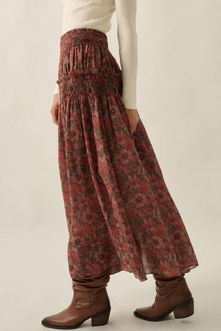 A person wearing a long, flowy floral maxi skirt in muted red and brown tones, paired with a cream top and brown ankle boots, shown against a plain light beige studio background.