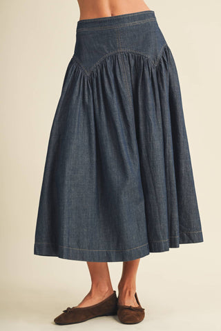 A dark blue denim midi skirt with a gathered waist and flared skirt, shown on a model wearing brown suede flats against a warm beige background.