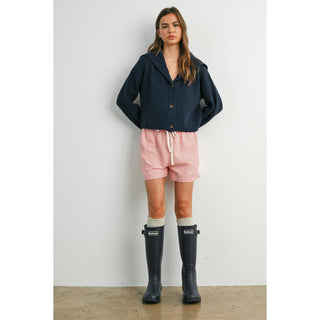 Woman wearing a navy cardigan, pink shorts, and black rain boots on a plain background