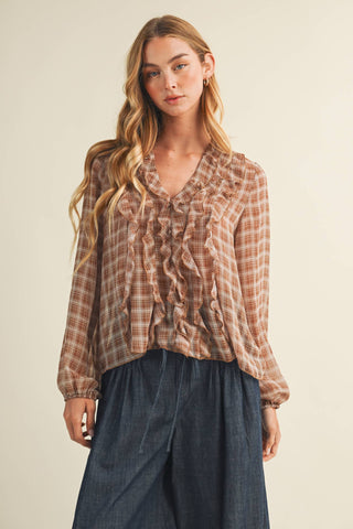 A woman modeling a long-sleeve ruffled blouse with a brown and beige plaid pattern, paired with dark blue wide-leg pants, against a plain beige background.