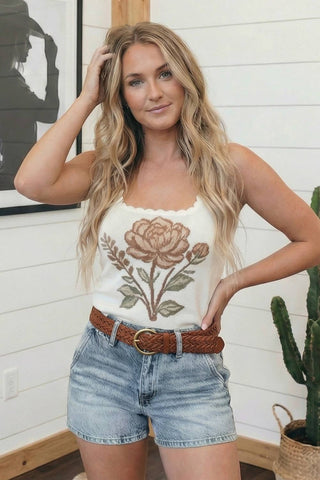 Woman wearing a white tank top with floral embroidery and denim shorts indoors.