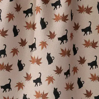 A close-up of a fabric surface with a repeating pattern of black cat silhouettes and brown/orange autumn leaves on a light beige background.