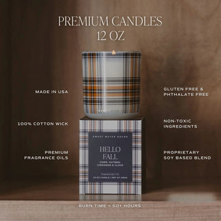 A premium 12 oz candle shown with its branded box on a wooden surface against a warm brown background. The candle has a plaid pattern in brown, beige, and black, with a lit wick.