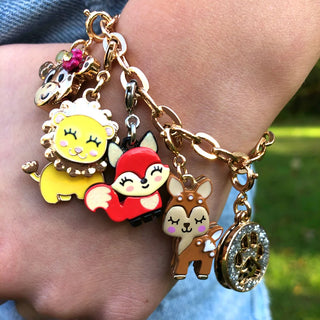 A close-up of a gold-tone charm bracelet worn on a wrist. The bracelet features multiple colorful enamel animal charms, including a yellow lion, a red fox, a brown deer, and a silver-toned locket charm, set against a blurred outdoor background.