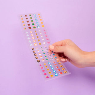 A hand holds a clear plastic sheet filled with many small colorful cartoon stickers against a light purple background.