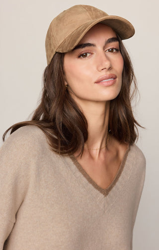 A woman wearing a tan baseball cap and a beige V-neck sweater, photographed against a plain light beige background.