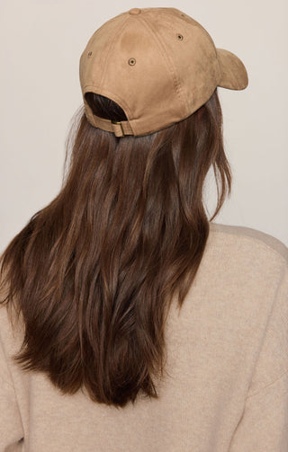 A person viewed from the back wearing a tan baseball cap with long brown hair and a light beige sweater against a plain light background.