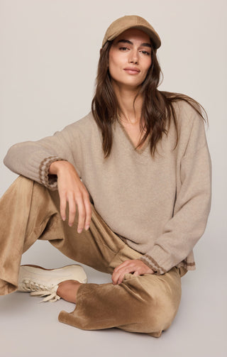 A woman sits cross-legged on a light gray studio floor wearing a beige knit V-neck sweater, matching beige cap, tan pants, and white sneakers.