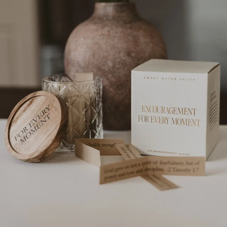 A boxed candle product labeled 'Sweet Water Decor' with the phrase 'Encouragement for Every Moment,' shown beside a wooden lid engraved 'For Every Moment' and a glass candle holder on a tabletop.