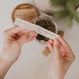 Hands holding a small paper strip with text, with a blurred background of a plant and wooden items.