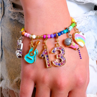A close-up of a person's wrist and hand wearing a colorful beaded charm bracelet. The bracelet features gold-tone metal rings and charms, including a turquoise guitar, a pink and purple rhinestone '13', a rainbow heart, a pink microphone, and pastel beads.