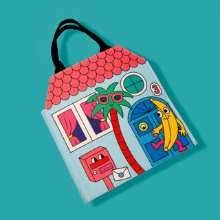 A light blue house-shaped tote bag with black handles, featuring a colorful cartoon illustration of a pink roof, blue door, red palm tree, and a yellow banana character on a teal background.