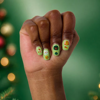 A close-up of a hand with light green Christmas-themed nail art, featuring festive illustrations like a Santa Claus, a snowman, a Christmas tree, and a gingerbread man, set against a blurred green holiday background.
