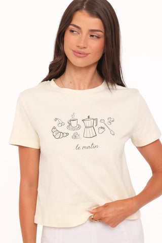 A woman wearing a cream-colored short-sleeve t-shirt with a black line art graphic of breakfast items and the French text 'le matin' on the front, set against a plain white background.