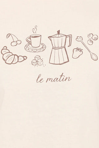 A close-up of a light beige fabric surface with a brown line-art breakfast illustration and the words 'le matin' printed below. Visible motifs include a croissant, cherries, a coffee cup on a saucer, a moka pot, a strawberry, a spoon, and sugar cubes.