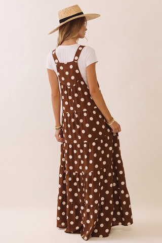 A woman wearing a brown and white polka dot overall maxi dress over a white t-shirt, paired with a straw hat, photographed from behind against a plain beige background.