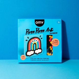 A boxed craft kit labeled 'omy Pom Pom Art RAINBOW' on a bright blue studio background. The package shows a rainbow-shaped pom pom canvas, a window with colorful pom pom beads, and orange callout stickers indicating 'FLUO NEON' and '+ 500 POMPOMS POMPOMS'.