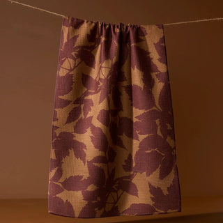 A single curtain panel hanging from a rope against a warm brown wall, featuring a textured waffle-weave pattern of dark plum purple and tan leaf motifs.