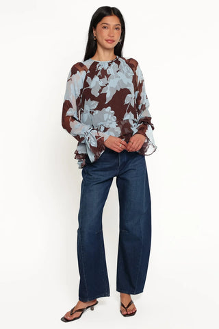 A woman modeling a sheer long-sleeve blouse with a brown and light blue floral pattern, paired with dark blue wide-leg jeans and black sandals, against a plain white studio background.