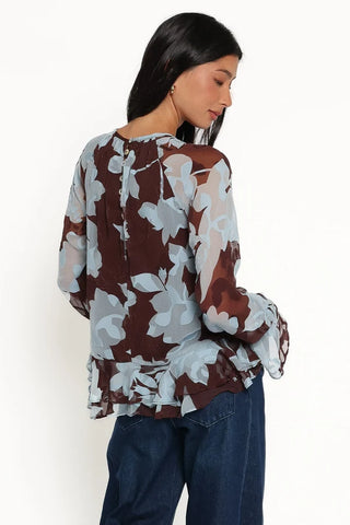 A woman wearing a sheer long-sleeve blouse with a brown, white, and light blue floral pattern, paired with dark blue jeans, shown from a three-quarters back view against a plain white background.