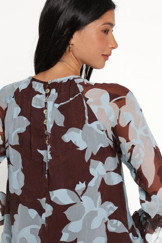 A close-up back view of a woman wearing a sheer floral blouse with a brown base and light blue-gray floral print, featuring a buttoned neckline.
