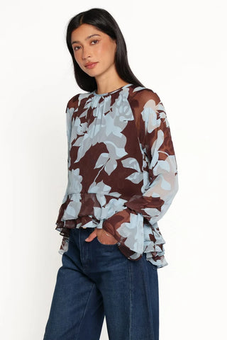 A woman modeling a sheer long-sleeve blouse with a brown and light blue floral pattern, paired with dark blue jeans, against a plain white studio background.