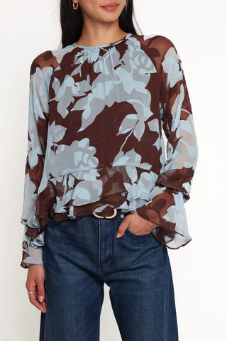 A woman wearing a sheer long-sleeve blouse with a brown, beige, and light blue floral pattern, tucked into dark blue jeans.